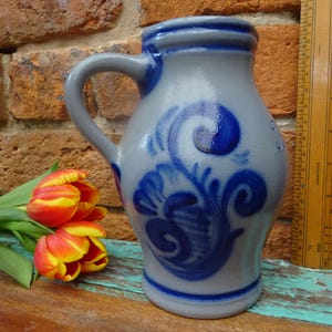 May include: A grey ceramic jug with a blue floral design and a handle. The jug is decorated with a blue floral pattern. A wooden ruler is next to the jug. A bouquet of red and yellow tulips rests on a wooden surface.