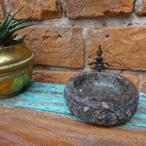 May include: A small, round, dark grey stone bowl with a detailed carving, topped with a small bronze figure. A brass and green planter with a succulent plant sits to the left. The background is a brick wall.