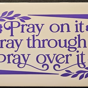 Könnte beinhalten: Eine rechteckige weiße Fliese mit den Worten "Pray on it, pray through it, pray over it" in lila Schrift. Der Text ist von floralen und Blattdesigns in der gleichen lila Farbe umgeben.