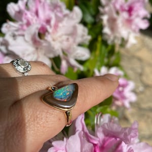 May include: A gold ring with a triangular opal gemstone set in a bezel setting. The opal has a rainbow of colors, including green, blue, and purple. The ring is on a finger against a background of pink flowers.