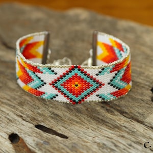 Pode incluir: Pulseira de punho com contas com um design nativo americano. A pulseira apresenta um fundo branco com padrões geométricos turquesa, vermelhos, laranja e amarelos. A pulseira tem cerca de 3,8 cm de largura.