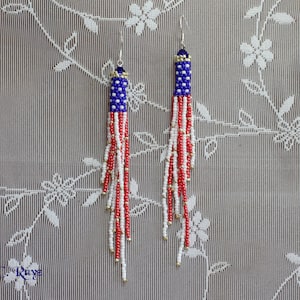 May include: Handmade beaded earrings in a patriotic design. The earrings feature a blue beaded top with red and white beaded strands, mimicking the American flag. Gold-colored beads accent the ends of the strands. The earrings hang from silver-colored hooks.