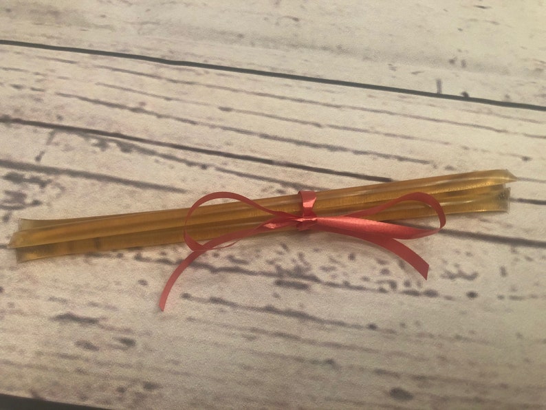 Puede incluir: Un conjunto de palitos de miel atados con una cinta roja. Los palitos, de color &aacute;mbar dorado, est&aacute;n dispuestos en paralelo. La cinta roja brillante a&ntilde;ade un toque decorativo, sugiriendo un regalo o una presentaci&oacute;n cuidada.