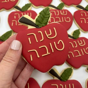 Peut inclure: Biscuits rouges en forme de pomme avec l'inscription hébraïque "Shanah Tovah" en glaçage beige. Chaque biscuit a une feuille verte et une tige marron. Les biscuits sont disposés sur une surface blanche.