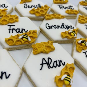 Puede incluir: Primer plano de galletas de azúcar decoradas. Cada galleta tiene forma hexagonal con glaseado blanco y un nombre escrito en negro. Están decoradas con panal de abeja amarillo y una pequeña abeja. Las galletas son de color marrón claro.