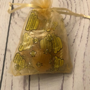 Puede incluir: Una peque&ntilde;a bolsa transparente de color amarillo dorado con cord&oacute;n, llena de peque&ntilde;os caramelos amarillos con sabor a miel. La bolsa est&aacute; impresa con texto amarillo y gr&aacute;ficos de abejas. El texto dice "Primrose Candy Co." y "Made with US Honey."
