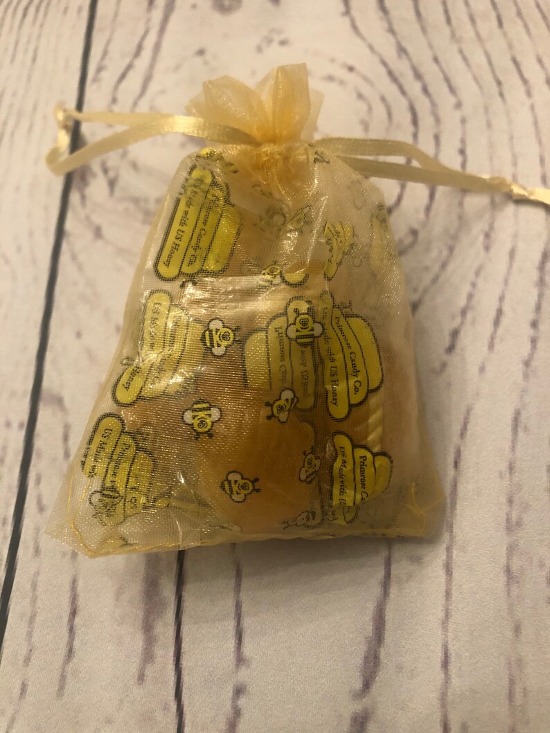 Puede incluir: Una peque&ntilde;a bolsa transparente de color amarillo dorado con cord&oacute;n, llena de peque&ntilde;os caramelos amarillos con sabor a miel. La bolsa est&aacute; impresa con texto amarillo y gr&aacute;ficos de abejas. El texto dice "Primrose Candy Co." y "Made with US Honey."