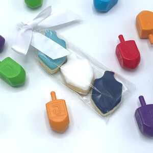 Puede incluir: Una bolsa de plástico transparente llena de tres galletas de azúcar decoradas con forma de trompo. Las galletas son azules, blancas y azul oscuro. La bolsa está atada con una cinta blanca. Hay varios trompos de plástico de colores esparcidos alrededor de la bolsa.