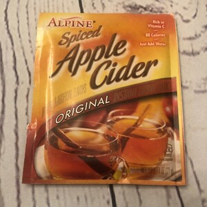 Puede incluir: Un paquete de mezcla de bebida instant&aacute;nea Alpine Spiced Apple Cider. El paquete es naranja y marr&oacute;n con las palabras "Spiced Apple Cider" en letras grandes. Presenta im&aacute;genes de dos tazas de sidra con palitos de canela. El paquete indica "Rich in Vitamin C".