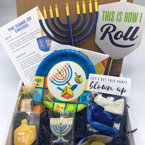 Puede incluir: Una caja blanca llena de artículos para fiestas de Hanukkah. La caja contiene una dreidel azul y blanca, una menorá azul y blanca, un plato de papel azul y blanco, un globo azul y blanco, una moneda de oro y una galleta azul y blanca. La caja también incluye un letrero que dice "Let's get this party blown up".