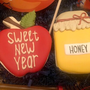 Puede incluir: Dos galletas decoradas: una manzana roja con "SWEET NEW YEAR" en blanco y un tarro amarillo con la etiqueta "HONEY". La manzana tiene una hoja verde y un tallo marr&oacute;n. El tarro tiene una etiqueta blanca y una tapa beige con un lazo rojo.