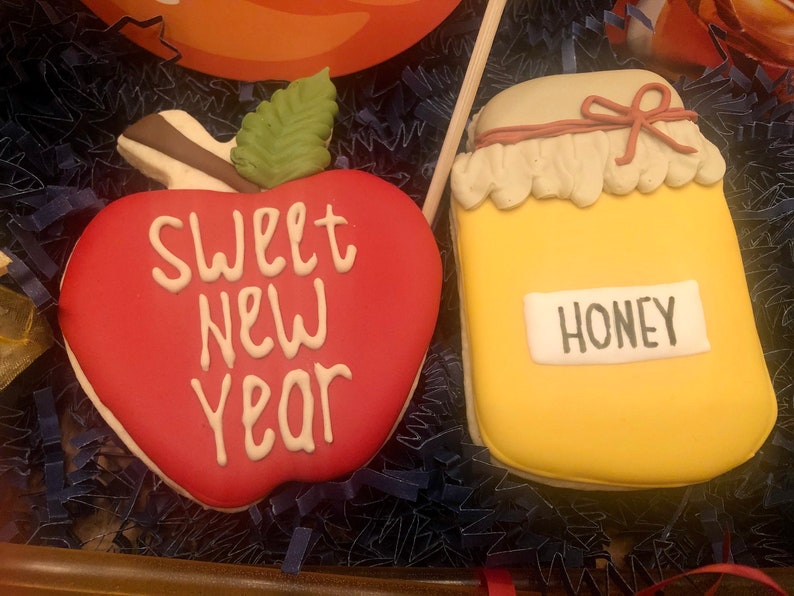 Puede incluir: Dos galletas decoradas: una manzana roja con "SWEET NEW YEAR" en blanco y un tarro amarillo con la etiqueta "HONEY". La manzana tiene una hoja verde y un tallo marr&oacute;n. El tarro tiene una etiqueta blanca y una tapa beige con un lazo rojo.