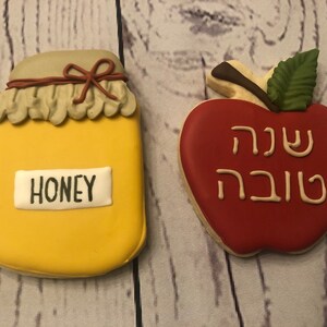 Puede incluir: Dos galletas decoradas: un tarro de miel amarillo con "HONEY" en una etiqueta blanca y una manzana roja con texto hebreo. El tarro de miel tiene un lazo marr&oacute;n y una tapa beige. La manzana tiene una hoja verde y un tallo marr&oacute;n.