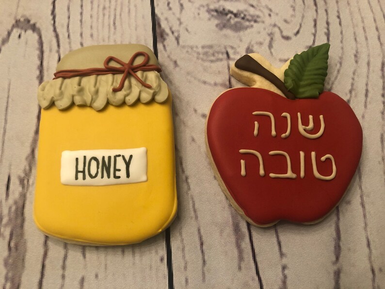 Puede incluir: Dos galletas decoradas: un tarro de miel amarillo con "HONEY" en una etiqueta blanca y una manzana roja con texto hebreo. El tarro de miel tiene un lazo marr&oacute;n y una tapa beige. La manzana tiene una hoja verde y un tallo marr&oacute;n.