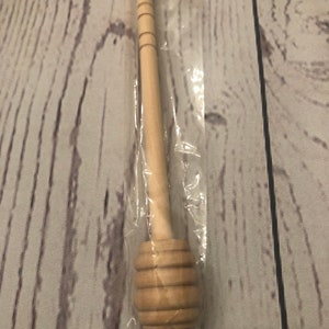 Puede incluir: Un cuchar&oacute;n de miel de madera, empaquetado en pl&aacute;stico transparente. El cuchar&oacute;n presenta un mango largo con anillos tallados y una cabeza redondeada y acanalada para recoger la miel. La madera tiene un acabado claro y natural.