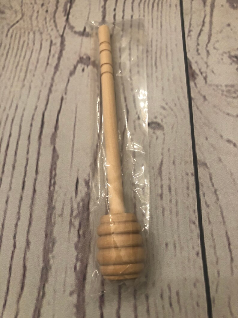 Puede incluir: Un cuchar&oacute;n de miel de madera, empaquetado en pl&aacute;stico transparente. El cuchar&oacute;n presenta un mango largo con anillos tallados y una cabeza redondeada y acanalada para recoger la miel. La madera tiene un acabado claro y natural.