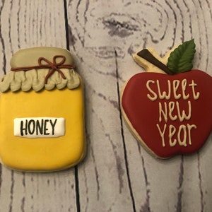Puede incluir: Dos galletas de az&uacute;car decoradas. Una tiene forma de tarro de miel con la palabra "HONEY". La otra es una galleta con forma de manzana con las palabras "SWEET NEW YEAR". Las galletas est&aacute;n sobre un fondo de madera.