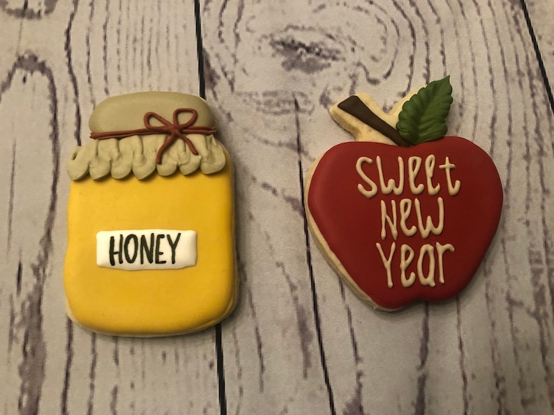 Puede incluir: Dos galletas de az&uacute;car decoradas. Una tiene forma de tarro de miel con la palabra "HONEY". La otra es una galleta con forma de manzana con las palabras "SWEET NEW YEAR". Las galletas est&aacute;n sobre un fondo de madera.
