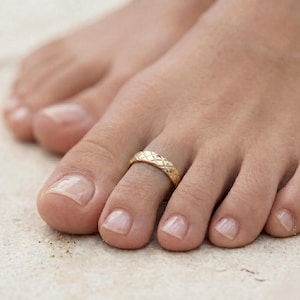 May include: A close-up of a foot with a gold-colored toe ring. The ring has a textured, quilted design. The toenails are neatly manicured. The background is a blurred, neutral tone.