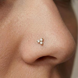 May include: A gold nose stud featuring three small, clear, round-cut gemstones arranged in a triangular cluster. The jewelry is set against the skin of a nose, highlighting the delicate design and sparkle of the gemstones.