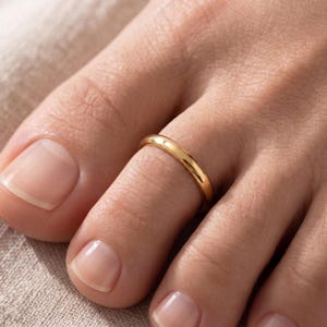 May include: A polished gold-tone metal toe ring with a smooth, rounded band design. The ring features a simple, continuous circular shape that sits securely around the second toe, showcasing a reflective, high-shine finish on the metal surface.