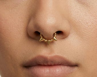 May include: Close-up of a gold septum ring featuring a series of small, pointed spikes. The jewelry is suspended from the septum and is centered below the nose. The skin tone is light, and the lips are a natural color.