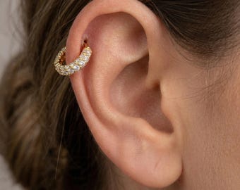 May include: A gold-colored, hoop-style earring with a row of small, clear gemstones. The earring is set in a gold metal and is worn on the upper ear. The image shows a close-up of the earring and part of the ear.