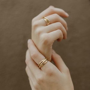 May include: Two gold-colored rings, each with a triple-band design, are worn on fingers. The rings have a smooth, polished finish. The background is a solid, warm brown color. The rings are a simple, elegant jewelry item.