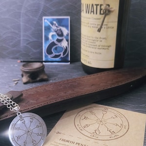 May include: A collection of esoteric items, including a silver pendant with Hebrew text, a brown leather sheath, a bottle with a label, and a card with a pentacle design and text. The items are arranged on a textured surface.