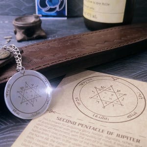 May include: A silver pendant with a chain, featuring Hebrew text and symbols, lies near a paper with similar symbols and text. A bottle with a label and a small decorative item are in the background. The paper reads "Second Pentacle of Jupiter."