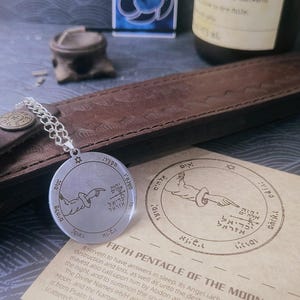 May include: A circular silver-toned metal pendant on a chain, engraved with a hand pointing toward Hebrew text and a star symbol. It rests on a paper card titled Fifth Pentacle of the Moon, which features matching illustrations and descriptive English text.