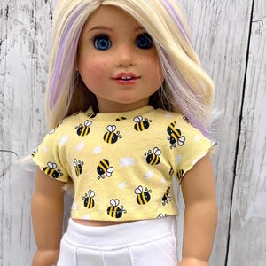 May include: A doll with blonde hair and blue eyes wearing a yellow crop top with a bee pattern and white pants. The crop top has a raw edge and features black and yellow bees and white cloud shapes.