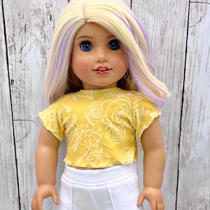 May include: A doll wearing a yellow crop top with a white floral pattern and white pants. The doll has blonde hair with purple streaks, blue eyes, and freckles. The doll is standing in front of a white wooden background.