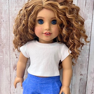 May include: A doll with curly blonde hair, blue eyes, and freckles, wearing a white t-shirt and blue sweatpants. The doll is posed against a light wood-grain background.