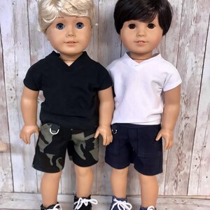 Może przedstawiać: Dwie lalki w swobodnych strojach. Lalka po lewej stronie ma blond włosy i nosi czarną koszulkę z kr&oacute;tkim rękawem, kr&oacute;tkie spodenki w kamuflaż i czarne buty sportowe. Lalka po prawej stronie ma ciemne włosy i nosi białą koszulkę z kr&oacute;tkim rękawem, kr&oacute;tkie spodenki granatowe i granatowe buty sportowe.
