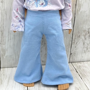 May include: A doll wearing a light blue corduroy pants with a flared leg design. The doll is also wearing a long-sleeved lavender shirt with a floral pattern on the sleeves and chest. The outfit is displayed on a light wood surface.