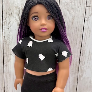 May include: A doll with long black hair featuring purple highlights, light skin, and pink eyes. The doll wears a black short-sleeved crop top with a white ghost pattern and a white crew neckline, paired with solid black leggings and purple lipstick.