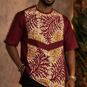 May include: A burgundy short-sleeved shirt with a patterned front panel. The panel features a cream and yellow leaf design. The shirt has a crew neck and is paired with black pants. A gold watch and rings are visible.