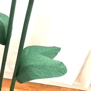 May include: Close-up of a green paper flower stem with two leaves. The leaves are made of crinkled green paper and are folded in a way that resembles a leaf.