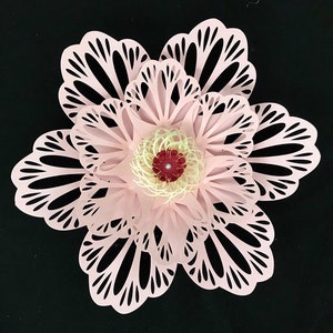 May include: A layered paper flower in shades of pink, with intricate cut-out petal designs. The center features a cream-colored spiral with a dark red accent and a pearl-like bead. The flower is set against a black background.