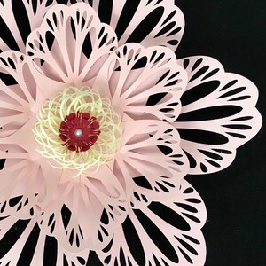 May include: A large, paper flower in shades of light pink, with intricate cut-out petal designs. The center features a cream-colored spiral and a dark red accent, set against a black background. A decorative floral element.