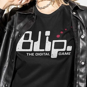 May include: Black t-shirt with the white text "Blip. THE DIGITAL GAME". The shirt is worn under a black leather jacket. A silver chain necklace is also visible.