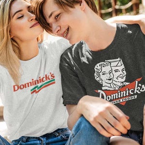May include: Two people wearing t-shirts with the "Dominick's" logo. One shirt is white with red and green text, the other is dark gray with a vintage diner graphic and the words "Dominick's Diner Foods".