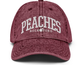 May include: A maroon baseball cap with the word "PEACHES" in white block letters arched across the front. Below the word "PEACHES" is the word "ROCKFORD" and a peach graphic. The cap has a curved brim.