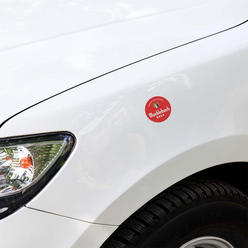 May include: A red and white round sticker with the text "Muehlebach BEER" is affixed to the white fender of a car. The sticker also features a small emblem and the phrase "ENJOY THE FINEST".