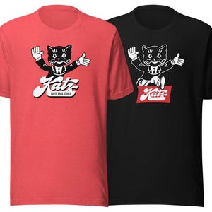 May include: Two t-shirts with a black and white illustration of a cat wearing a suit and holding a red sign that says "Katz Super Drug Stores". The t-shirt on the left is red and the t-shirt on the right is black.
