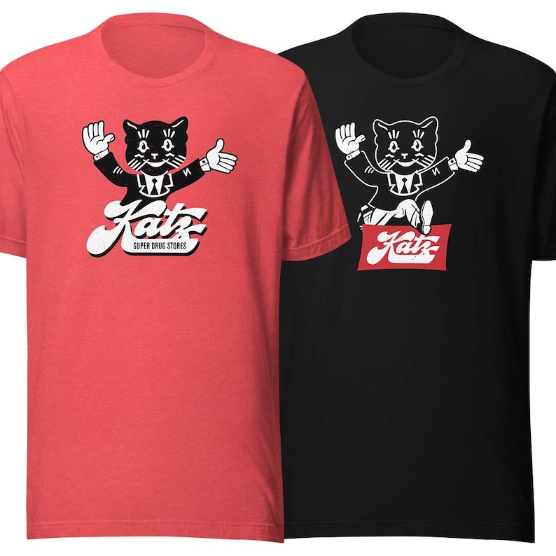 May include: Two t-shirts with a black and white illustration of a cat wearing a suit and holding a red sign that says "Katz Super Drug Stores". The t-shirt on the left is red and the t-shirt on the right is black.