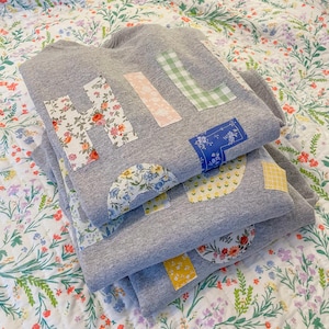 May include: Three grey sweatshirts are stacked on a floral patterned bedspread. Each sweatshirt has the word "Hi, Hello" appliquéd on the front with fabric patches in various floral and patterned designs. The sweatshirts are folded neatly.