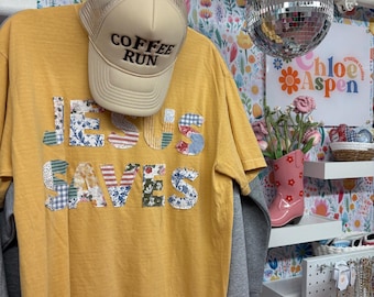 May include: Mustard yellow t-shirt with patchwork letters spelling "JESUS SAVES". A beige baseball cap with "COFFEE RUN" is on top. The background features disco balls and floral wallpaper.
