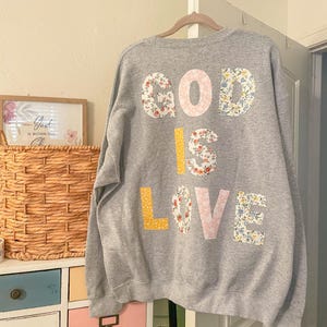 May include: A grey sweatshirt with the words "GOD IS LOVE" appliquéd on the back. The letters are in a mix of floral patterns and solid colours, including pink, yellow, and white. The sweatshirt is hanging on a wooden hanger.